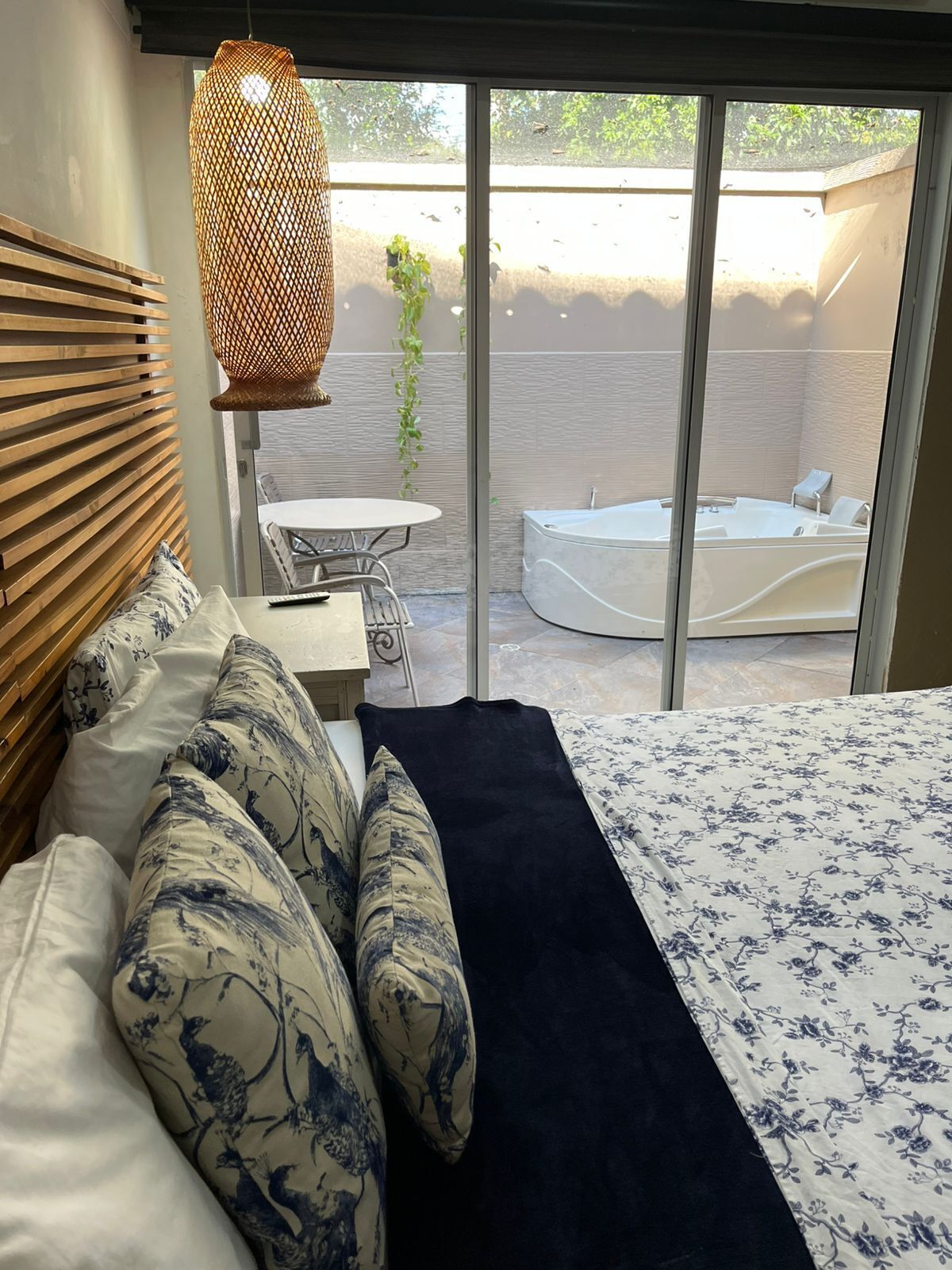 Un dormitorio con cama, mesita de noche, lámpara colgante y vistas a un área exterior con jacuzzi y mesa.