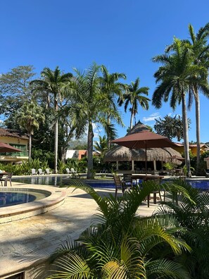 Ein Poolbereich eines Resorts mit Palmen, einer Gazebo mit Reetdach und Liegestühlen.