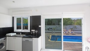 A modern kitchen with a washing machine, a dishwasher, and a large window overlooking a swimming pool area. - Protaras Holiday Villa Gd44 (Paralimni)