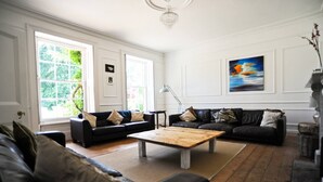 A spacious living room with two black leather sofas, a wooden coffee table, and a large window providing natural light. - Manor House Moreton - Entire Holiday Home (Dorchester)