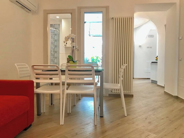 A modern living room with a dining area, a red sofa, white chairs, and a view of the kitchen through an open door. - Trifoglio Apartment in S Pietro (Maiori)