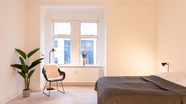 A bedroom with a bed, a chair, a potted plant, and a window with a view of buildings. - Charming House Close to Everything (Aarhus)