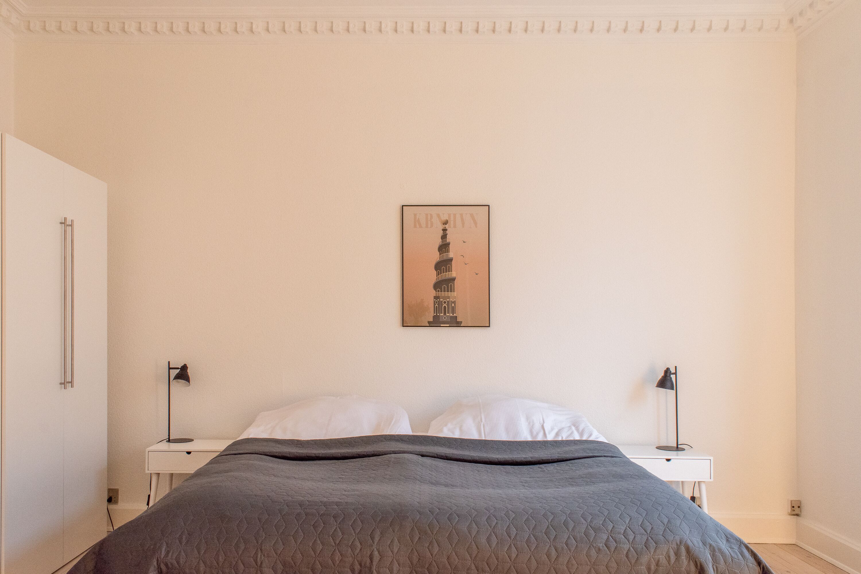 A bedroom with a bed, two bedside tables, a wall-mounted picture, and a white door.