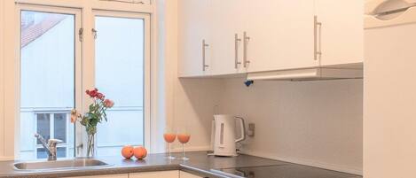 A modern kitchen with white cabinets, a black and white checkered floor, a window with flowers, and a kettle on the counter.
