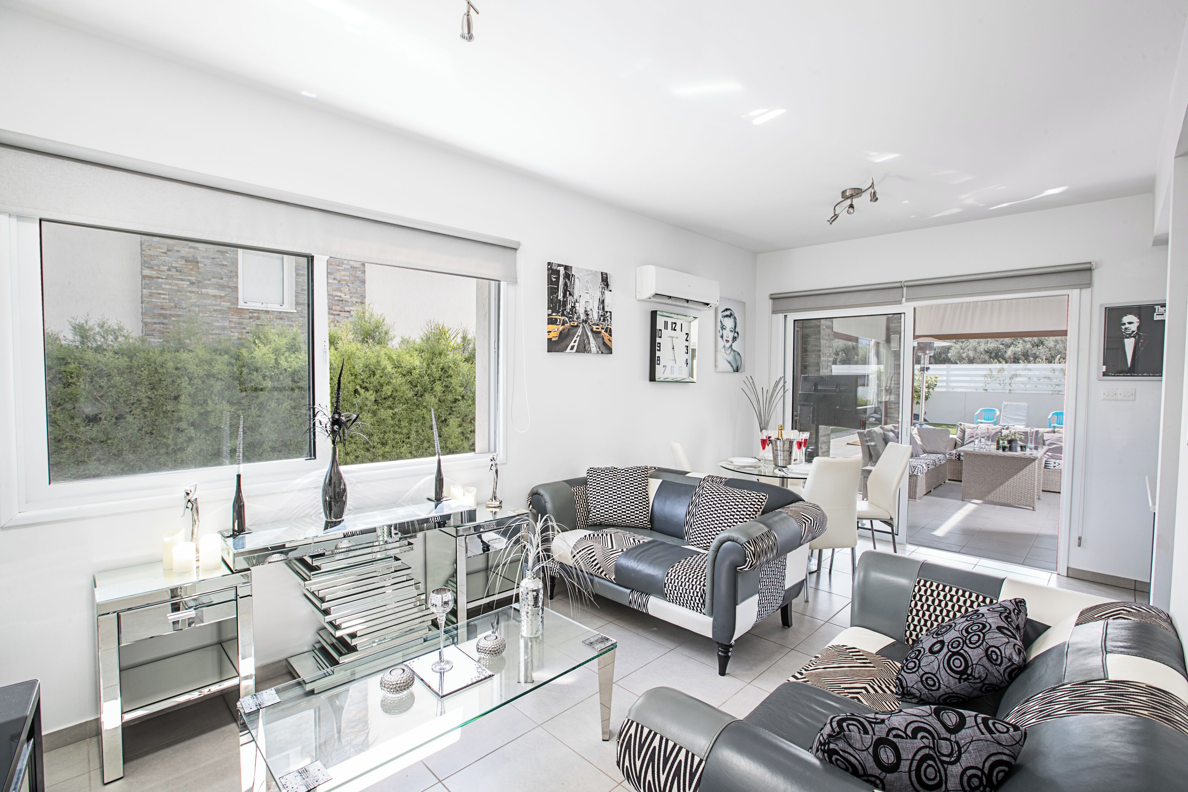 A modern living room with a glass coffee table, a grey sofa, and a dining area with outdoor seating.