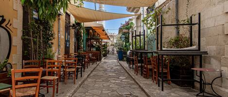 A narrow cobblestone street with outdoor seating, wooden chairs, and tables under shaded areas.