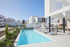 Un área de piscina moderna con sillones de descanso y sombrillas, rodeada de edificios con balcones y un cielo despejado.