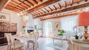 A spacious living area with a wooden ceiling, a dining table set with chairs, a fireplace, and a large window with curtains. - Elisa Farmhouse Apartment in Wine Resort (Lucca)