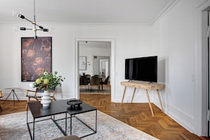 A modern living room with a wooden herringbone floor, a black TV on a wooden stand, a white vase with flowers, and a large abstract painting on the wall. - Square - Spacious 6-bdr Apt Near Nyhavn (Copenhagen)