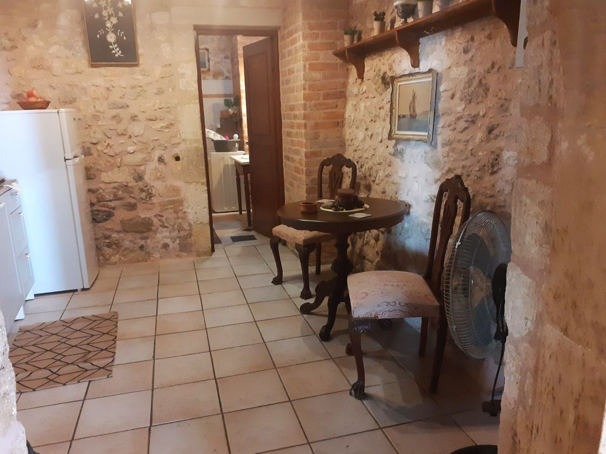 A kitchen with a tiled floor, a wooden dining table with chairs, a white refrigerator, and a fan.