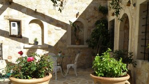 Une cour bordée de murs en pierre, deux grands pots en terre cuite contenant des plantes, une chaise en métal blanche et une petite table.