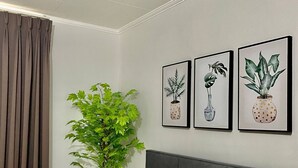A neatly made bed with a grey headboard, a large green plant, and three framed pictures of plants on the wall. - 1 Bedroom With Free Parking (Morong)