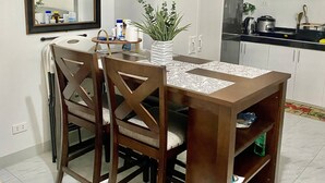 A compact kitchen with white cabinets, a dining table with chairs, a clock on the wall, and a potted plant. - 1 Bedroom With Free Parking (Morong)