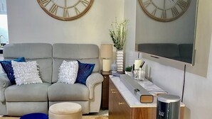 A living room with a grey sofa, two round wall clocks, a coffee table, ottoman, and a TV on a wooden cabinet. - 1 Bedroom With Free Parking (Morong)