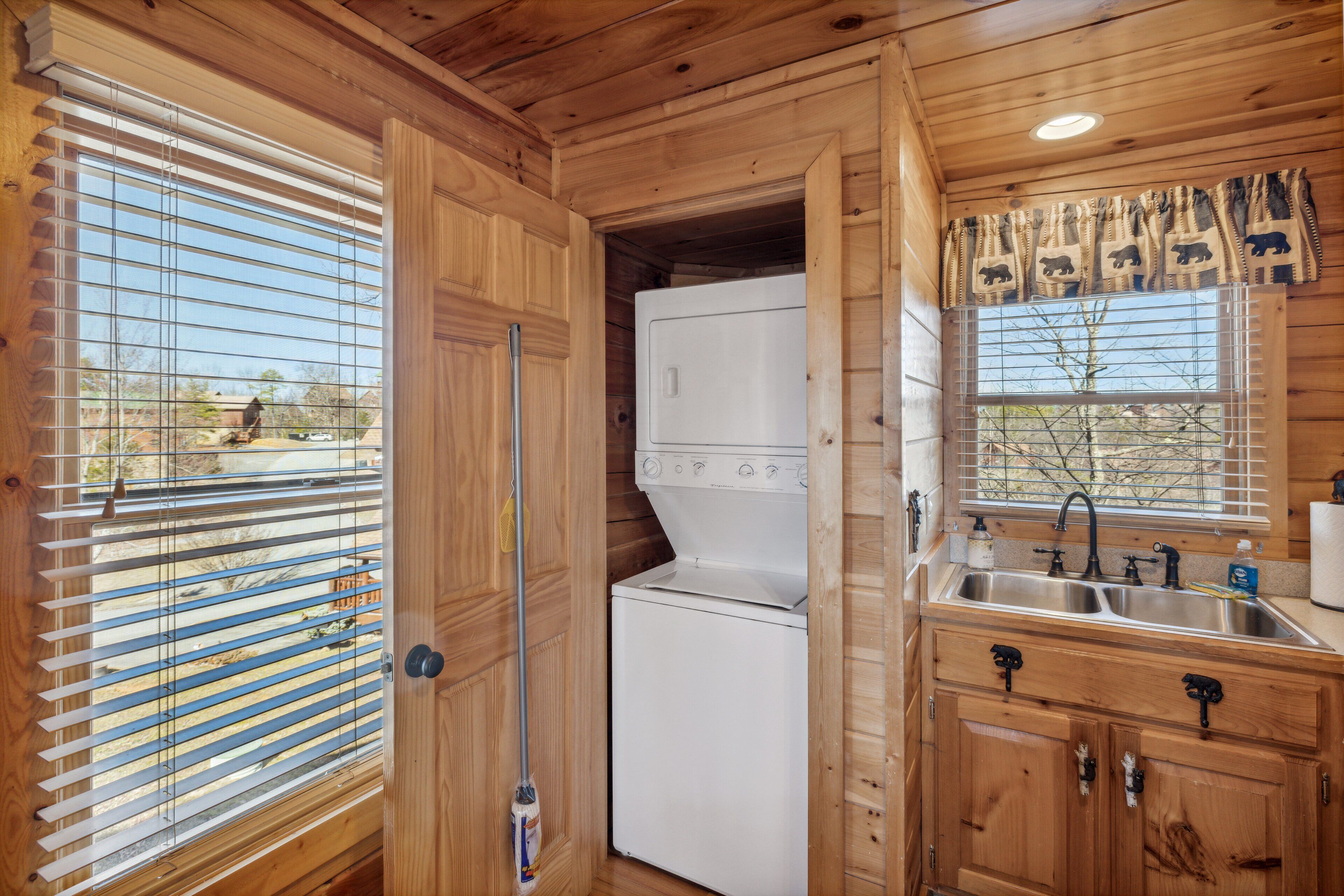 A compact kitchen with wooden cabinetry, a built-in washer and dryer, a sink, and a window with blinds.