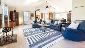 Un vaste salon avec un canapé bleu foncé, une table basse en bois et un tapis à motifs. On y trouve également un coin repas avec une table et des chaises en bois, et une cuisine à l’arrière-plan.
