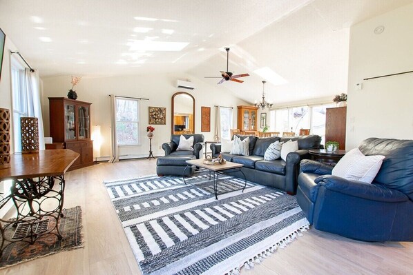 A spacious living room with a dark blue sofa, a wooden coffee table, and a patterned rug. There is a dining area with a wooden table and chairs, and a kitchen in the background. - Large Home w/ Fire Pit and Dogs Welcome! (Chatham)