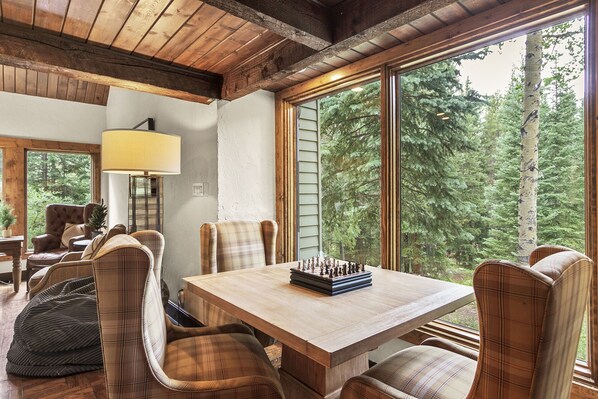 A cozy dining area with a wooden table and plaid chairs, a chessboard, and a view of a forest through large windows.