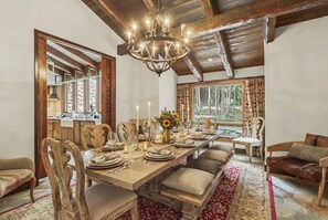 Une salle à manger avec une longue table dressée pour six personnes, des chaises en bois, un lustre et une vue sur la forêt par la fenêtre.