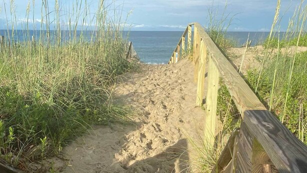 Ein hölzerner Steg führt zu einem Strand mit Sand und hohem Gras an beiden Seiten.