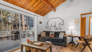 Un salon chaleureux avec une grande fenêtre, un plafond en bois, une décoration murale représentant un ours, un canapé, une table basse et une petite table avec une lampe.
