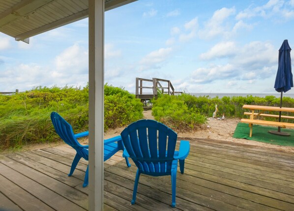 A wooden deck with blue chairs and a picnic table, overlooking a beach with a wooden structure in the distance. - Oceanfront Escape—Cape Cod Bay & Sandbars (East Sandwich)