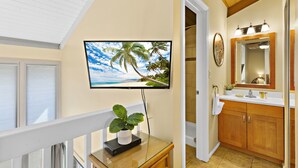 A modern bathroom with a large TV mounted on the wall, wooden cabinets, and a glass table with a plant. - Sugar Beach beauty, Your 2bd Oceanfront Escape (Kihei)