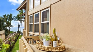 A wooden table with a woven placemat, a wine bottle, and a glass on a balcony overlooking a garden with palm trees and a view of the sea. - Sugar Beach beauty, Your 2bd Oceanfront Escape (Kihei)