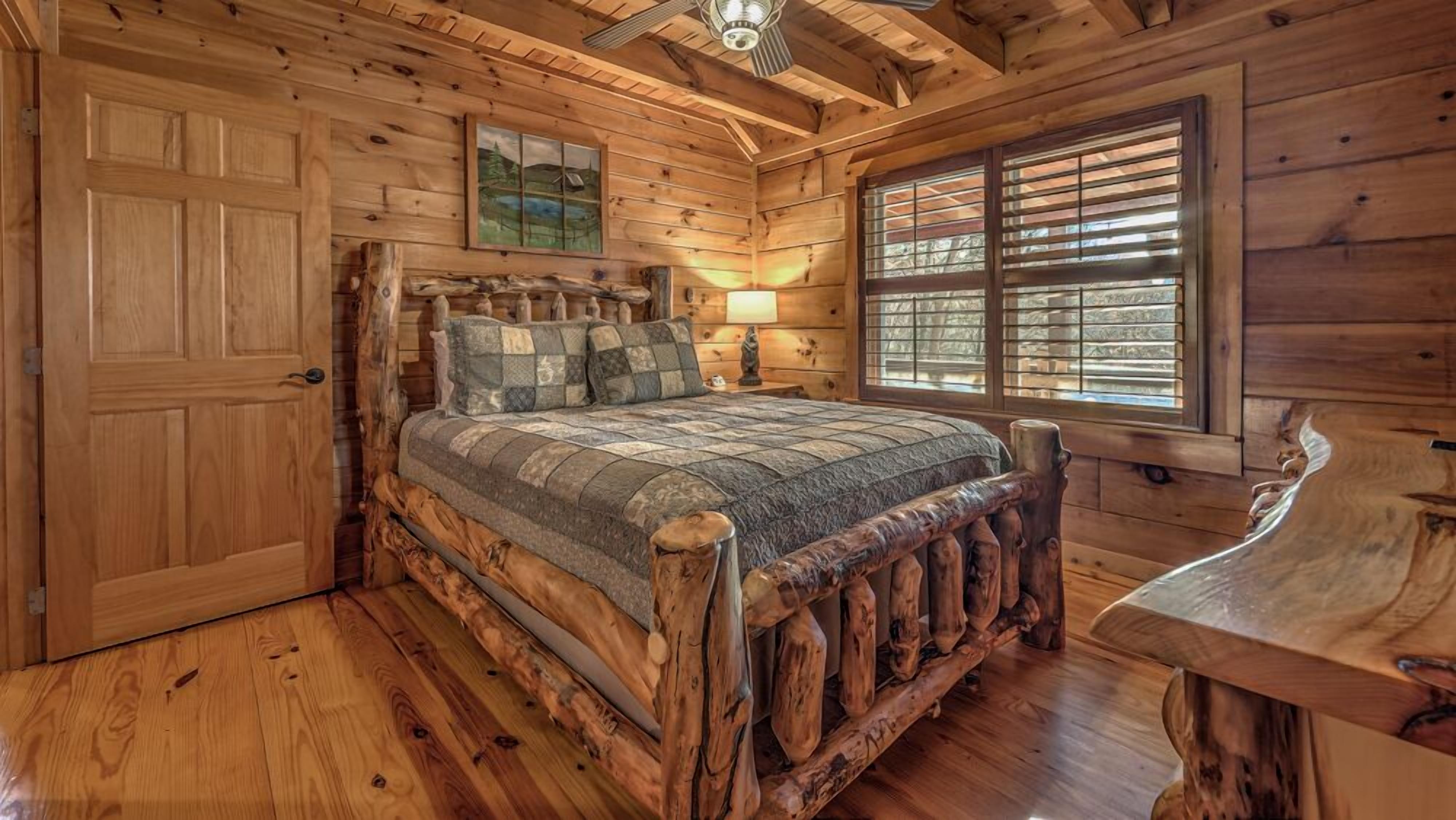 A log cabin bedroom with a wooden bed, a nightstand, a lamp, and a window with blinds.