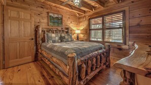 A log cabin bedroom with a wooden bed, a nightstand, a lamp, and a window with blinds. - Treehouse Hideaway - Pet Friendly | Hot Tub | Game Room (Cherry Log)