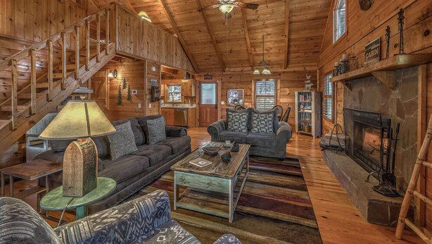 A log cabin interior with a staircase, two sofas, a coffee table, a fireplace, and a bookshelf. - Treehouse Hideaway - Pet Friendly | Hot Tub | Game Room (Cherry Log)
