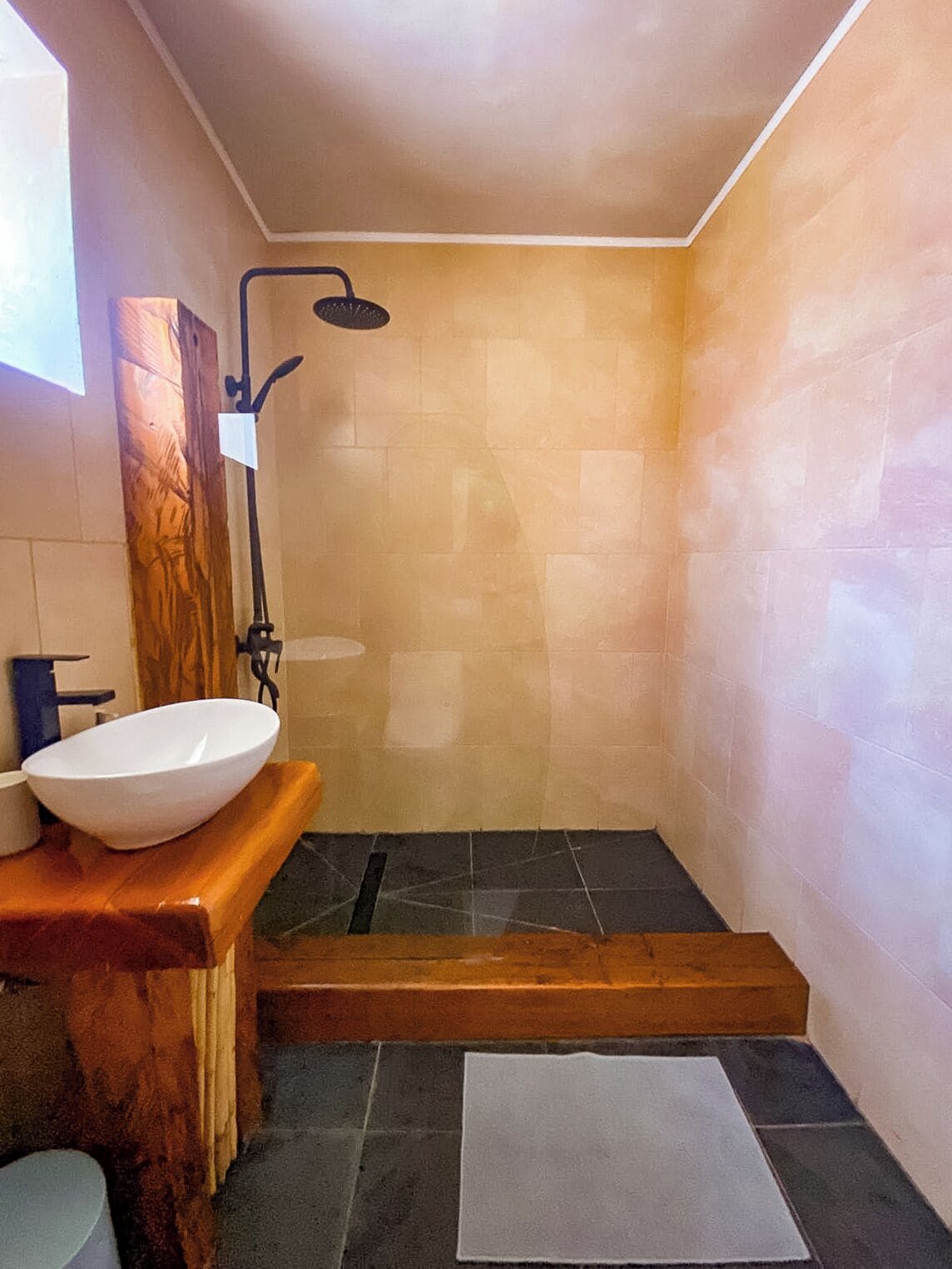 Une salle de bain avec un meuble-lavabo en bois, un lavabo blanc, une douche avec un tuyau noir et un sol carrelé.