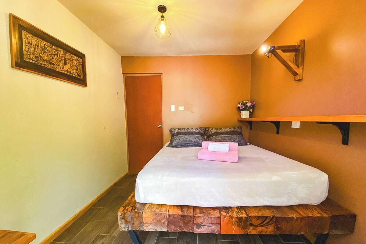 A bedroom with a bed on a wooden platform, a door, a wall-mounted shelf with a vase, and a framed picture on the wall.