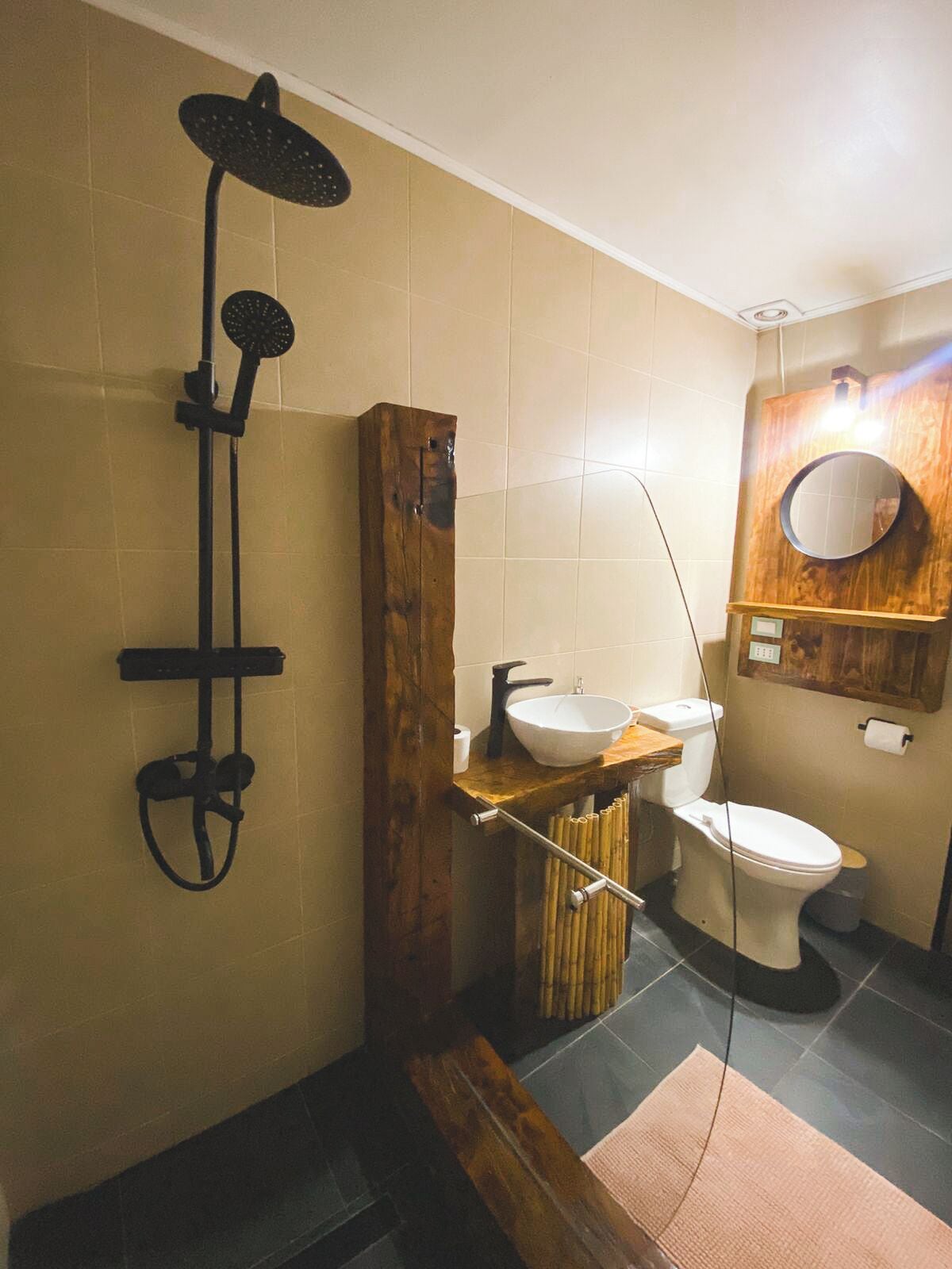 Un baño con tocador de madera, lavamanos blanco, inodoro y ducha con cabezal de ducha tipo lluvia.