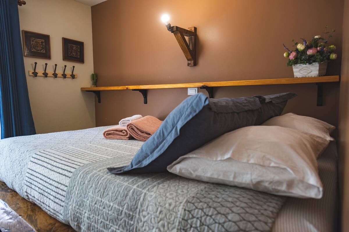 A neatly made bed with a patterned bedspread, a folded towel, and a pillow. There is a wooden shelf above the bed with a potted plant and a wall-mounted light fixture.