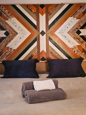 Una cama con dos almohadones oscuros y una toalla doblada, frente a una pared con un diseño geométrico.