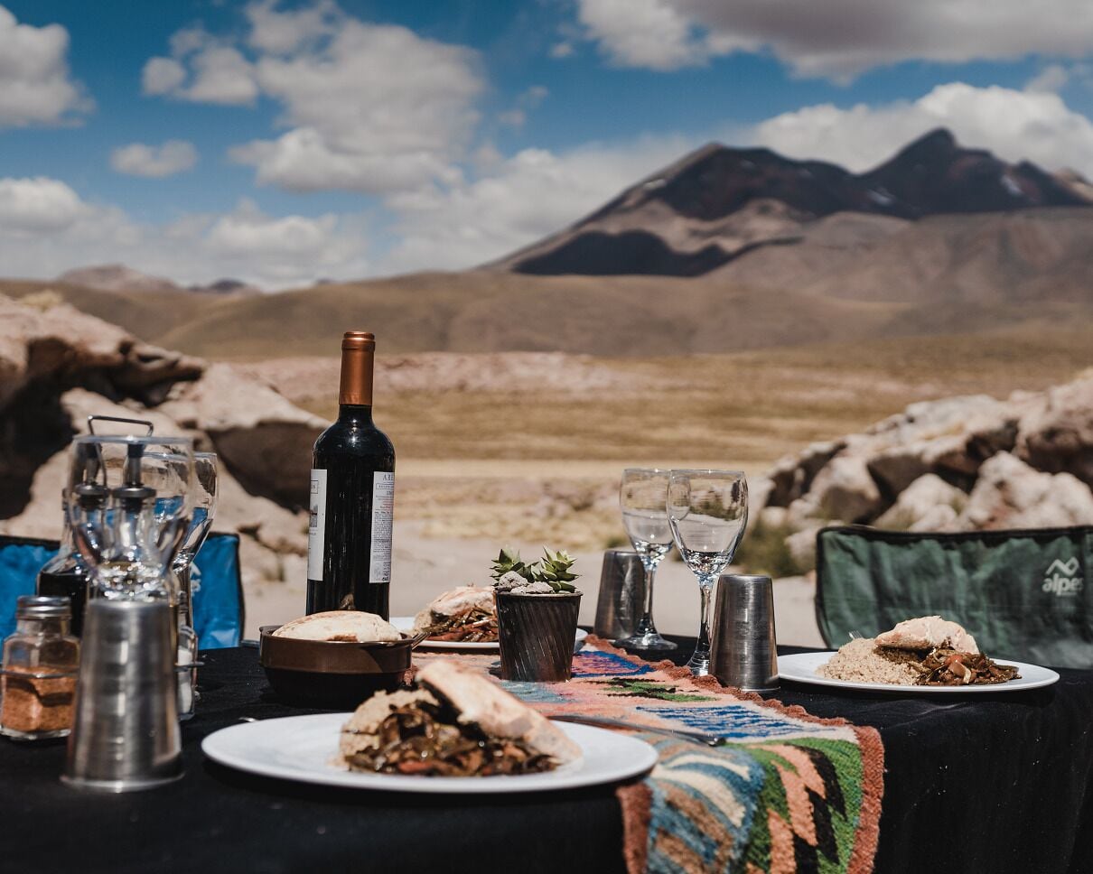 Mesa de comedor al aire libre con una botella de vino, copas y platos de comida, con montañas al fondo.