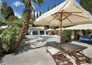 A pool area with lounge chairs and umbrellas, surrounded by palm trees and a building in the background.