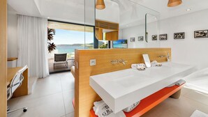 A modern bathroom with a large white sink, wooden vanity, and a large mirror. There is a view of the ocean through the glass door. - Villa Grace | Ocean View - Located in  Fabulous Anse des Cayes with Private Pool (Anse des Cayes)