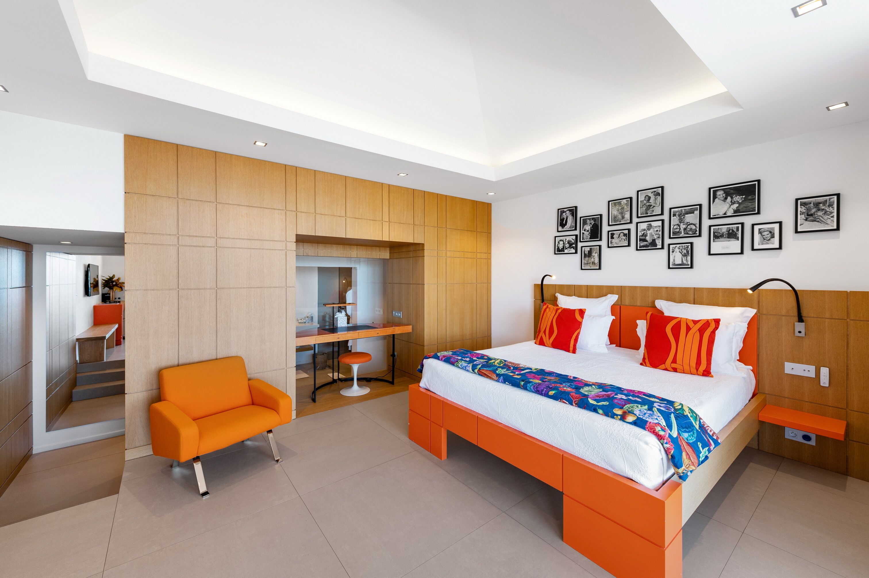 Une chambre moderne avec un grand lit, un fauteuil orange, un bureau et un mur décoré de tableaux encadrés.