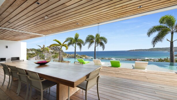 Un espace repas extérieur moderne, avec une table et des chaises en bois, offrant une vue sur une piscine et l’océan.