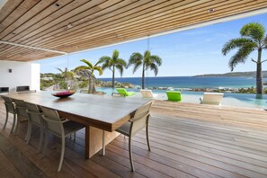 Un área de comedor exterior moderna con una mesa y sillas de madera, con vistas a una piscina y al océano.