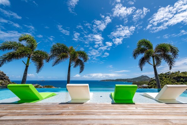 A poolside area with lounge chairs and palm trees, overlooking a clear blue sea and distant land.