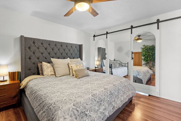 A bedroom with a tufted headboard, a large bed with patterned bedding, a nightstand, a mirror, and a ceiling fan. - Golden Chestnut, Cal King Bed, Sleeps 4, Centrally Located (Loveland)