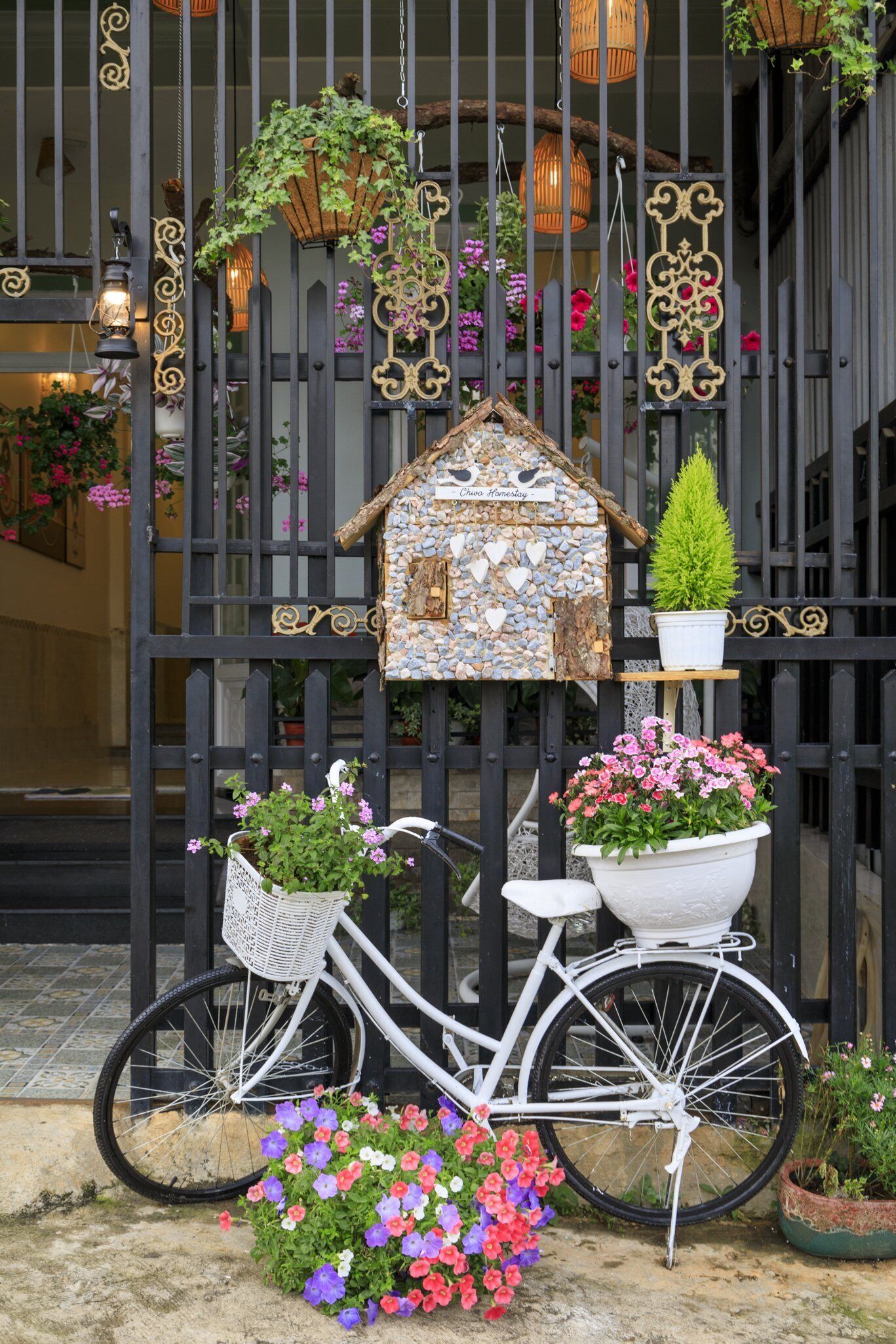 Un vélo blanc avec un panier et une petite décoration en forme de maison, le tout placé sur une clôture en métal noir.