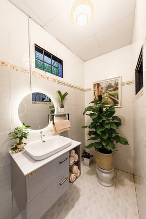 Een badkamer met een wit wastafelmeubel, een ronde spiegel, een plant in pot en een ingelijst schilderij aan de muur.