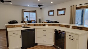 Une cuisine moderne avec des meubles blancs, un lave-vaisselle et un four encastré. Elle est dotée de deux fenêtres à cadre en bois et d’un ventilateur de plafond.