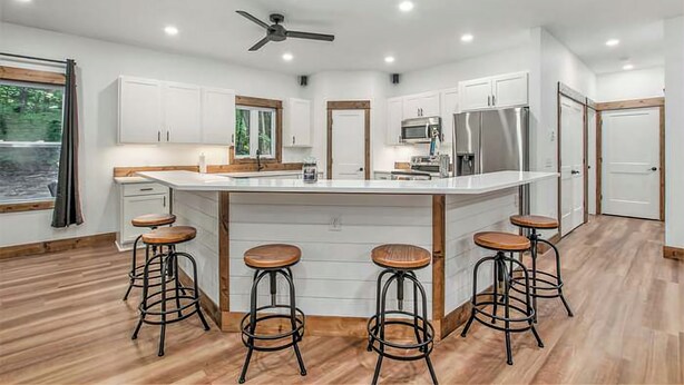 Une cuisine moderne avec des armoires blanches, un grand îlot central doté de tabourets, des appareils électroménagers en acier inoxydable et un ventilateur de plafond.