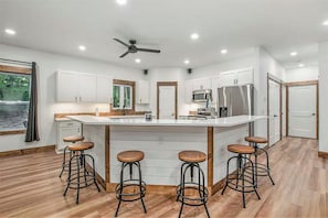 A modern kitchen with white cabinets, a large island with stools, stainless steel appliances, and a ceiling fan.