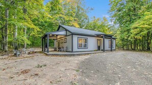 Une cabane dotée d’un porche couvert, d’une petite terrasse et d’une allée en gravier, le tout entouré d’arbres.